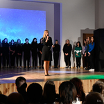 Nauczycielka zabiera głos na uroczystości, w tle: stoją uczniowie na scenie