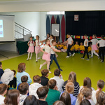 Młodzi uczestnicy spotkania tańczą w parach, chłopcy ubrani są w eleganckie ciuchy, dziewczynki mają na sobie białe bluzki oraz rajstopy oraz różowe spódniczki
