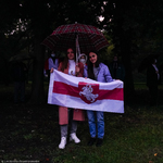 Dwie kobiety stojące pod parasolem w trakcie koncertu, trzymające biało-czerwono-białą flagę Białorusi