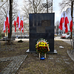 Wieniec i zapalony znicz pod pomnikiem Wielkiej Synagogi w otoczeniu biało-czerwonych flag