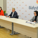 Uczestnicy konferencji siedzący przy stole na tle białej ścianki z logiem Miasta Białystok
