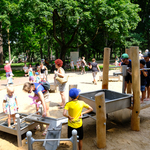 Dzieci bawią się na wodnym placu zabaw, w tle: rodzice i opiekunowie