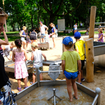 Dzieci bawią się na wodnym placu zabaw, w tle: rodzice i opiekunowie