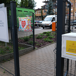 Biała skrzynka z żółtą nalepką na środki higieniczne zamontowana na ogrodzeniu budynku Centrum Aktywności Społecznej