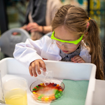Dziewczynka w okularach ochronnych, ubrana w fartuch wykonująca doświadczenie laboratoryjne