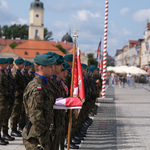 Żołnierze stojący w dwóch szeregach na placu, na pierwszym planie poczet ze sztandarem. W tle widać budynek ratusza i kamienice