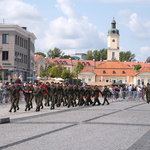 Żołnierze trójkami maszerujący przez Plac Jana Pawła II, pierwsi niosą złożoną flagę Polski, w tle widać ratusz i kamienice