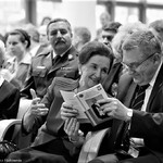 Uśmiechnięta Karolina Kaczorowska nachylająca się do Prezydenta Tadeusza Truskolaskiego, który ogląda książkę o mężu Karoliny - Ryszardzie Kaczorowskim, siedzą wśród innych gości honorowych podczas uroczystości, fotografia czarnobiała