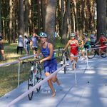 Zawodnicy triathlonu prowadzą rowery trasą biegnącą przez sosnowy las, w tle widzowie oglądający wyścig