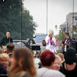 Grający na scenie zespół, na środku stoi śpiewająca blondwłosa wokalistka, w pierwszym planie widać tańczących nieostrych ludzi