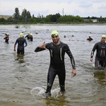 Uśmiechnięty zawodnik wychodzi z wody pokazując kciuk do góry, za nim kolejni uczestnicy wyścigu wychodzący z zalewu