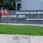 Mur z napisem Pomnik Obrońców Miasta, przed nim flagi Polski w stojakach