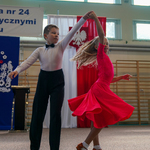 Chłopiec w koszuli oraz dzewiczynka w czerwonej sukience dają pokaz tańca towarzyskiego na parkiecie sali sportowej