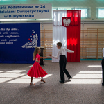 Chłopiec w koszuli oraz dzewiczynka w czerwonej sukience dają pokaz tańca towarzyskiego na parkiecie sali sportowej