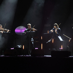 Koncert, na scenie czterech mężczyzn grających na instrumentach smyczkowych