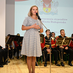 Radna Joanna Misiuk przemawia do mikrofonu ze sceny, za nią siedzi orkiestra