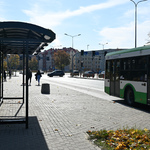 Po lewej stronie kadru zielony przystanek, po prawej odjeżdżający autobus, w tle bloki
