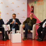 Czterech mężczyzn siedzących w fotelach podczas konferencji, drugi z lewej strony przemawia do mikrofonu