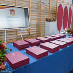 Czerwone teczki z listami gratulacyjnymi leżące na niebieskim stole, po lewej stronie naręcze róż, w tle duży ekran z prezentacją