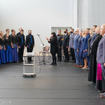 Po lewej śpiewający młodzieżowy chór w niebiesko-czarnych strojach dyrygowany przez czarnowłosą kobietę, po prawej stojący goście honorowi uroczystości