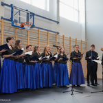 Młodzieżowy chór w niebiesko-czarnych strojach śpiewa dyrygowany przez kobietę