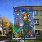 Szeroki kadr na mural na ścianie budynku Szkoły Podstawowej nr 22, po lewej widać rosnące wokół szkoły drzewa. Przed muralem stoi pomnik Marii Konopnickiej