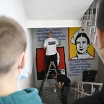 Dzieci stojące na schodach w szkole, przyglądające się jak na półpiętrze artyści malują na ścianie mural