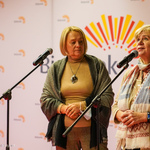 Dwie kobiety stoją przy mikrofonie, jedna o czymś opowiada