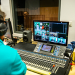 Studio nagrań, przed ekranem komputera siedzi młody mężczyzna w słuchawkach, zza ramienia widać na ekranie otwarte okna z obrazami z kilku kamer