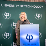 Przez mikrofon przemawia blondwłosa kobieta we wzorzystej, brązowo-czarej sukience