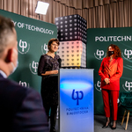 Przy mównicy przemawia kobieta w średnim wieku w czarnej sukience w kwiaty
