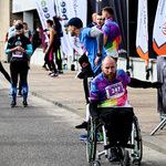 Mężczyzna poruszający się na wózku inwalidzkim ubrany w sportowe ubranie z numerem 247 przygotowuje się do startu, w tle: inni uczestnicy wydarzenia