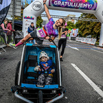 Uśmiechnięta kobieta wraz z chłopcem, w wózku podczas zakończenia biegu w tle: dmuchana brama mety
