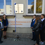 Uczniowie stoją przy pamiątkowej tablicy, na chodniku położono znicz, białą różę i kamienie