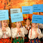 Dzieci ubrane w ludowe stroje trzymają w dłoniach tabliczki z napisami w języku polskim oraz ukraińskim