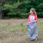 Dziewczynka podczas sprzątania ziemi trzyma w rękach szary worek na odpady zmieszane, w tle: las