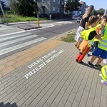 Na zdjęciu widać napis na chodniku, w tle dzieci w kamizelkach odblaskowych