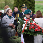 Młody przedstawiciel służb mundurowych trzyma wieniec kwiatowy, w tle inni uczestnicy uroczystości