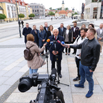 Widok z nad kamery na Prezydenta Miasta Białegostoku, który przemawia do mikrofonów trzymanych przez dziennikarzy. Na drugim planie radni oraz urzędnicy