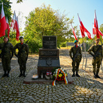 Mundurowi stoją na baczność obok pomnika, w tle ustawione flagi Polski