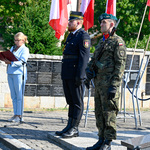 Prowadząca uroczystość mowi do mikrofonu, obok stoi dwóch mundurowych na baczność