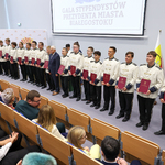 Prezydent stoi na scenie auli w otoczeniu uczniów ubranych w garnitury orkiestrowe 
