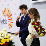 Uczniowie prowadzący uroczystość stoją przy mównicy, uczeń mówi do mikrofonu, uczennica trzyma bukiet kwiatów 