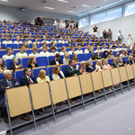 Urzędnicy, nauczyciele, uczniowie i inni goście siedzą na krzesłach dla publiczności w auli