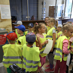 Dzieci w żółtych odblaskowych kamizelkach stoją obok urny do głosowania, opiekunka dzieci podaje im kartki, w tle stoi człowiek przebrany za maskotkę Budżetu Obywatelskiego 