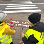 Uczniowie rozdają przechodniom odblaski, w tle malowany napis na przejściu dla pieszych 