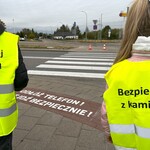 Uczniowie rozdają przechodniom odblaski, w tle malowany napis na przejściu dla pieszych 