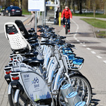 Rowery Biker stoją przy stojaku, w tle rowerzysta jedzie ścieżką rowerową