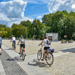 Białostoczanie jadą na rowerach przez Rynek Kościuszki w stronę ulicy Legionowej, w tle zielone drzewa oraz lekko zachmurzone błękitne niebo