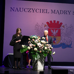 Prezydent Tadeusz Truskolaski stoi na scenie, obok kobieta mówi do mikrofonu, w tle grafika z herbem miasta 